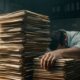 An overwhelmed, heavy-set man sits face-down at a desk behind towering stacks of files in a dimly lit office.