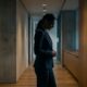 A professional woman in a dark business suit standing in a dimly lit office hallway, looking down with a somber expression to represent the emotional impact of workplace discrimination and retaliation.